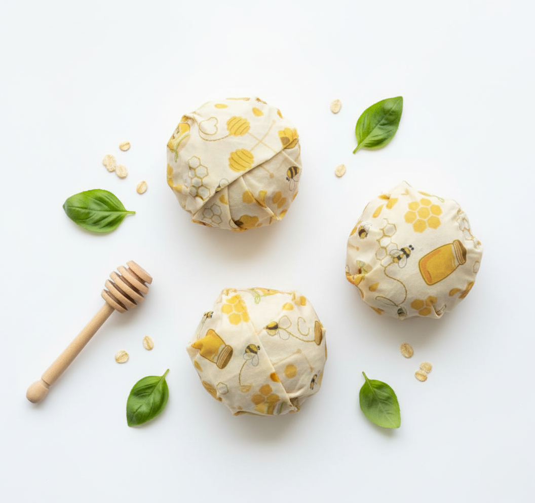 Three wrapped items in beeswax wraps on a white background with honeycomb pattern, basil leaves, and a honey dipper.